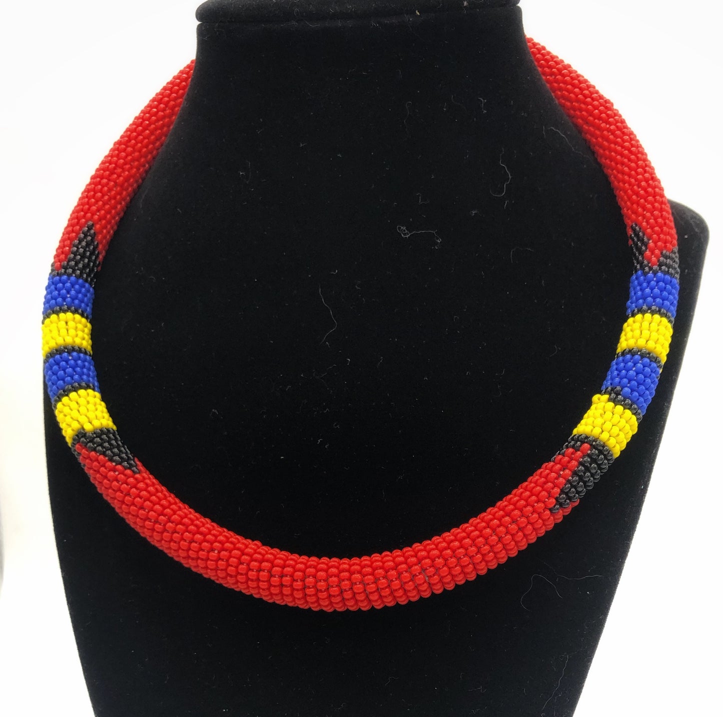 Wear the epitome of Kenya style with The Bintiah necklace. This necklace is handmade using colorful seed beads that are intricately and tightly wrapped around a solid piece fo make a stunning statement with any outfit. Red with blue, yellow and black accent beads