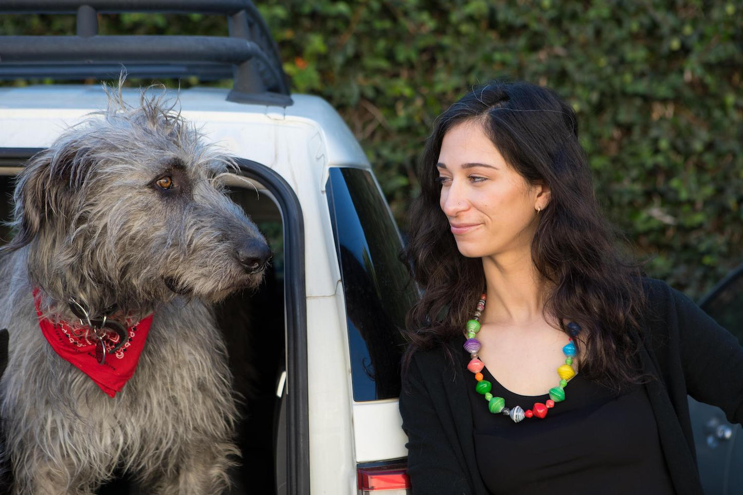 Woman wearing paper bead necklace looking at dog sitting in back of SUV with ivy in the background on the wall