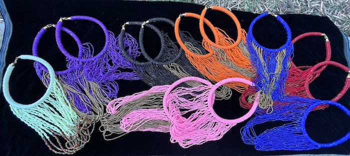 13 necklaces laid out on a black velvet cloth on the ground. Chocker style necklace with long strands of beads. Loops down and back up to chocker to connect. Cream, purple, black, orang, red, pink and royal blue solid and with gold beads shown.