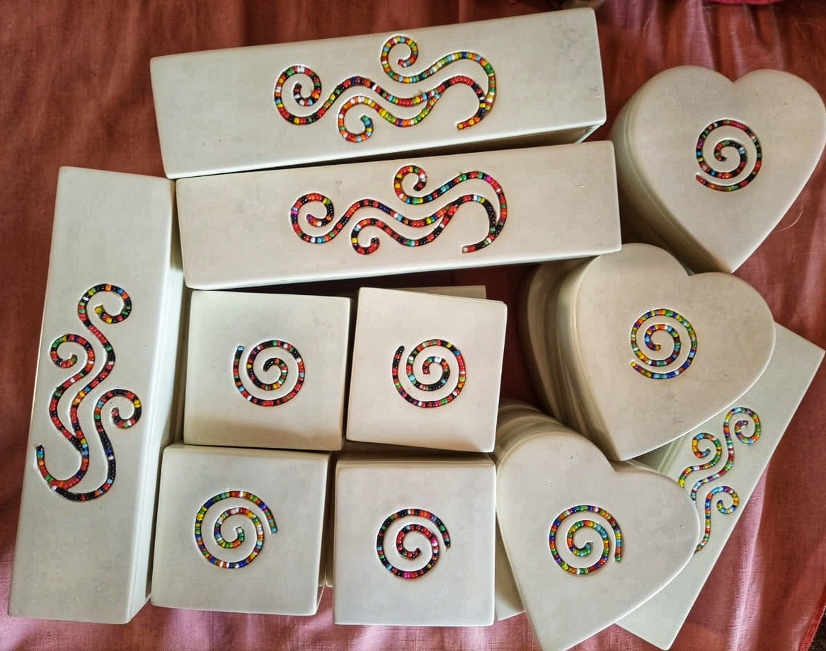 Soapstone trinket boxes 4 rectangular boxes, 4 square boxes, 3 heart shaped boxes stacked on top of more of the same. Rectangular box to the side of the hears on rose colored fabric.