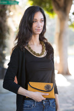 Load image into Gallery viewer, Young lady pictured from the hips up with black shirt, gold necklace and gold mini clutch purse and tree faded on the background