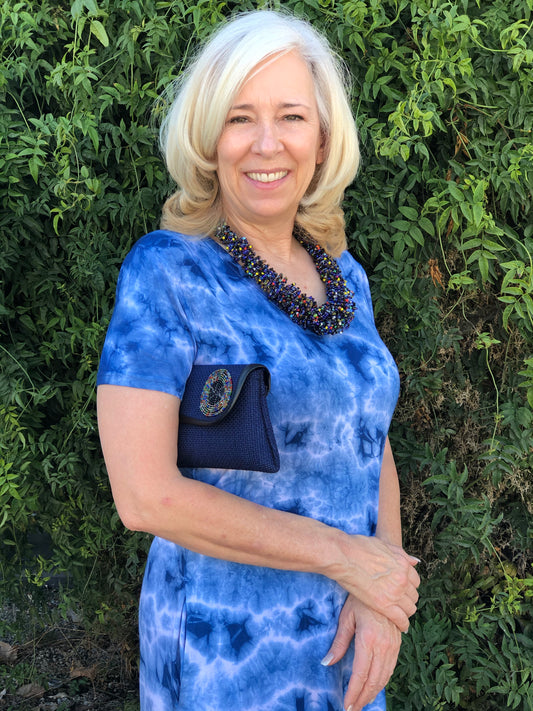 Woman in blue dress with platinum hair wearing a beaded necklace and holding mini clutch purse under arm