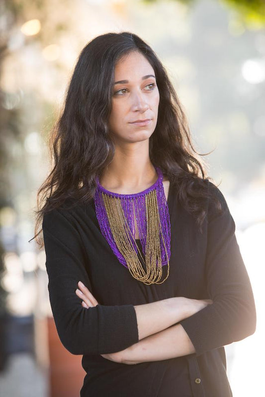 Model from waist up in black with long dark brown hair wearing Mary beaded purple and gold necklace. Chocker style necklace with long strands of purple and gold beads hanging down to breast area.  Loops down and back up to chocker to connect. 